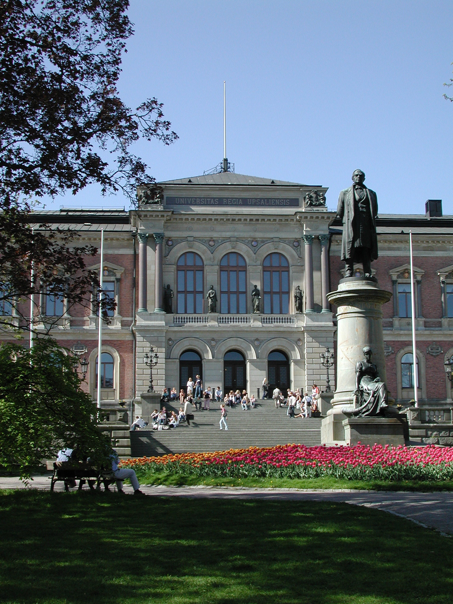 The image shows the grand facade of a university building, featuring large arched windows and a prominent entrance with steps leading up to it. In front of the building, there is a statue of a historical figure, surrounded by colorful flower beds. People are gathered on the steps and in the surrounding area, enjoying the day under a clear blue sky. Trees are partially framing the scene, enhancing the natural beauty of the campus.