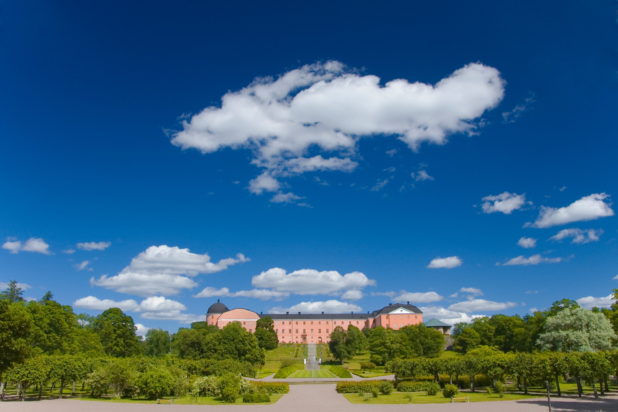 A scenic view of the Uppsala castle surrounded by lush greenery and trees, with a clear blue sky filled with fluffy white clouds overhead. The building features a domed roof and is set against a backdrop of vibrant foliage, with a pathway leading up to it.